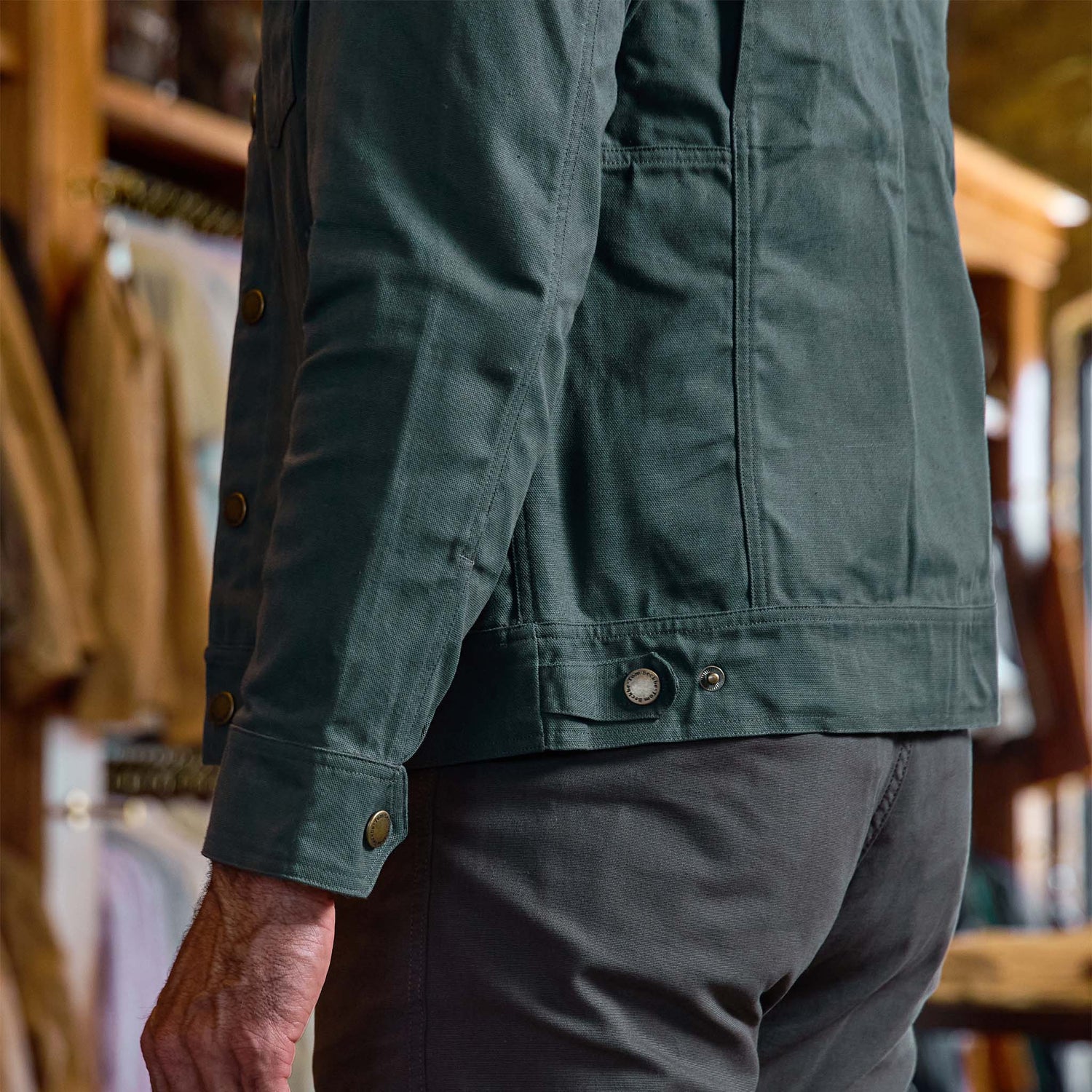 A man wearing the Tom Beckbe Crawford Jacket and gray pants stands indoors with clothing racks behind him.
