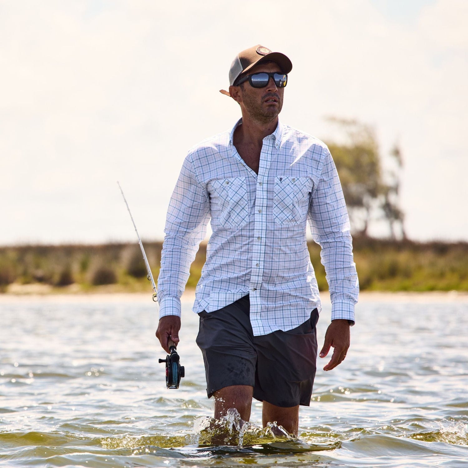 A man in Tom Beckbes Crenshaw Performance Shorts wades through waist-deep water, fishing under a partly cloudy sky.