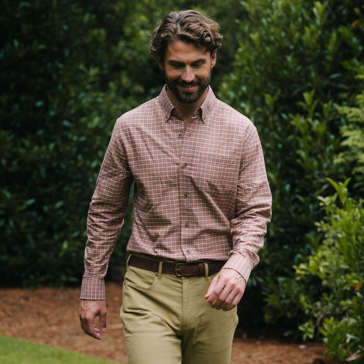 Man in a Tom Beckbe Culver Oxford Shirt and khakis strolls outside, surrounded by greenery and fresh air.