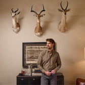 A man wears Tom Beckbe Reid Straight Leg Cotton Chino Pants in a room with antelope heads above a book and bottle cabinet.