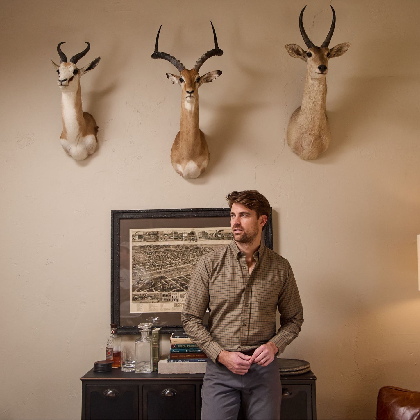 A man wears Tom Beckbe Reid Straight Leg Cotton Chino Pants in a room with antelope heads above a book and bottle cabinet.