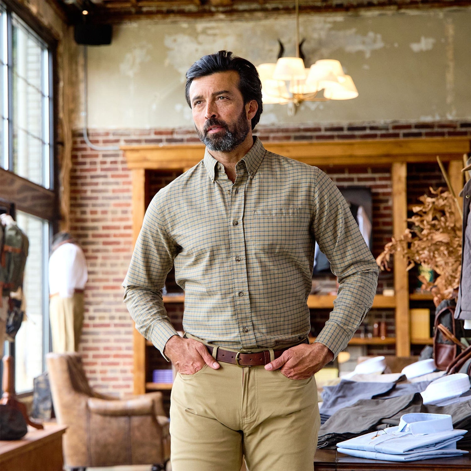 A man in a Tom Beckbe Culver Oxford Shirt and khaki pants stands in a store with brick walls and wooden shelves.