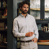 Man wearing the Tom Beckbe Culver Oxford Shirt, holding a glass in a room with shelves and glassware.