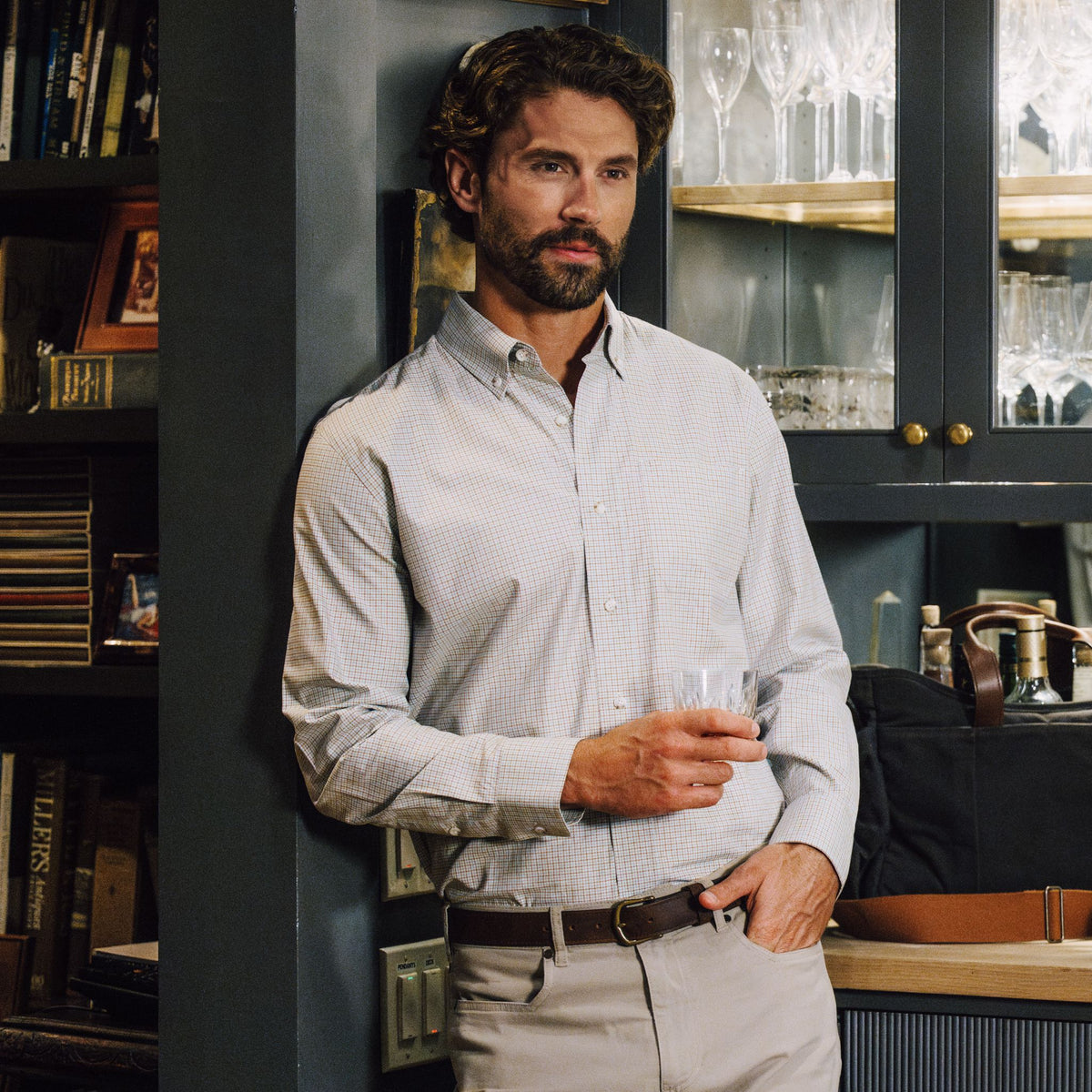 Man wearing the Tom Beckbe Culver Oxford Shirt, holding a glass in a room with shelves and glassware.