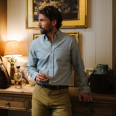 Man in a Tom Beckbe Culver Oxford Shirt and khakis holds a drink by a wooden desk in warm lighting.