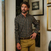 Man with a beard wearing a checkered shirt and khaki pants stands indoors by a wall with framed pictures.