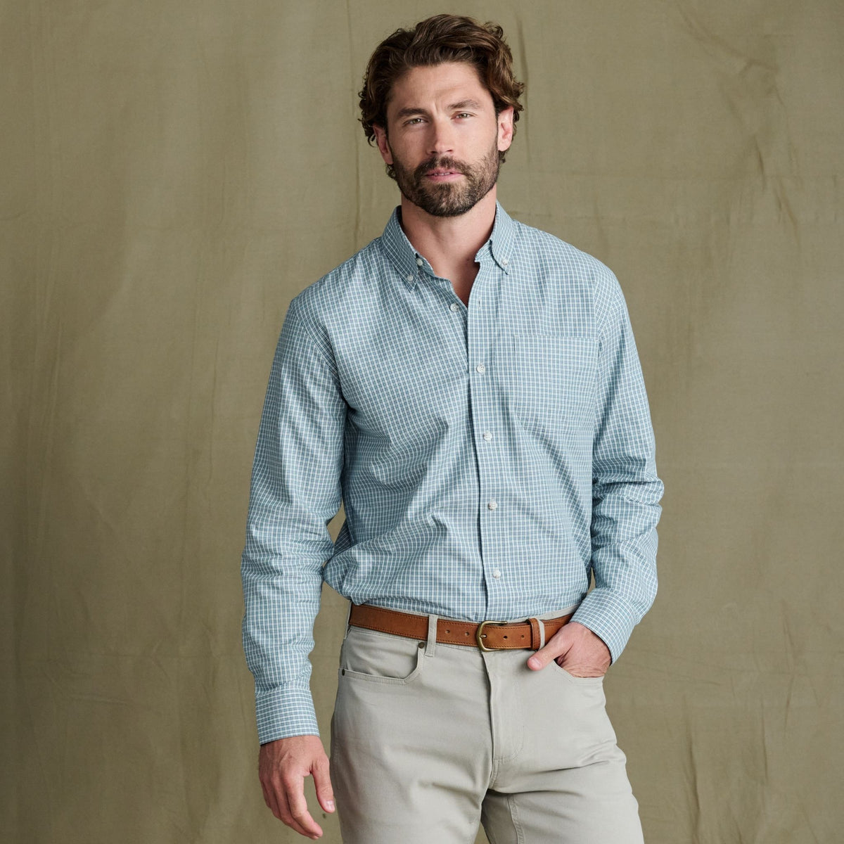 Man in a Tom Beckbe Ensley Pinpoint Oxford Shirt, beige pants, and brown belt, standing by a tan background.