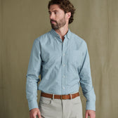 Man wears Tom Beckbe Ensley Pinpoint Oxford Shirt in light blue check over beige pants, brown belt; plain backdrop.