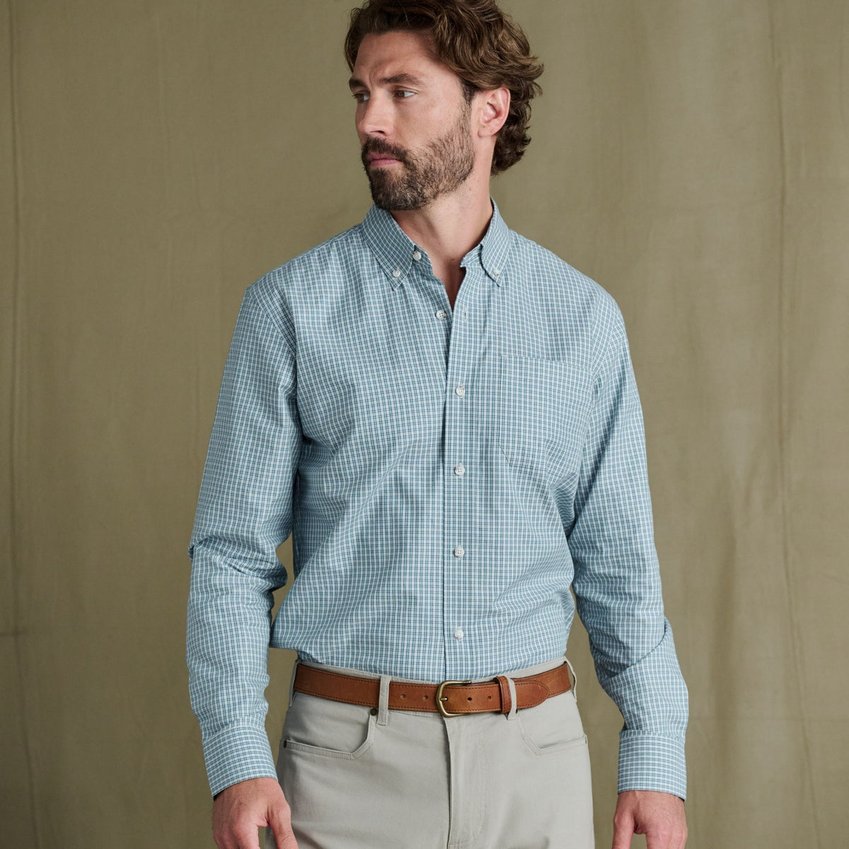 Man wears Tom Beckbe Ensley Pinpoint Oxford Shirt in light blue check over beige pants, brown belt; plain backdrop.