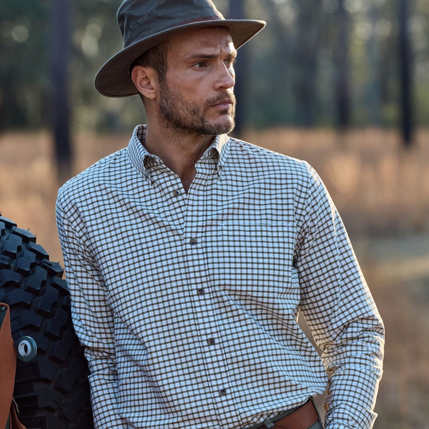A man in a Tom Beckbe Field Hat and checkered shirt stands outdoors by a large tire, looking sideways. Sparse trees behind.