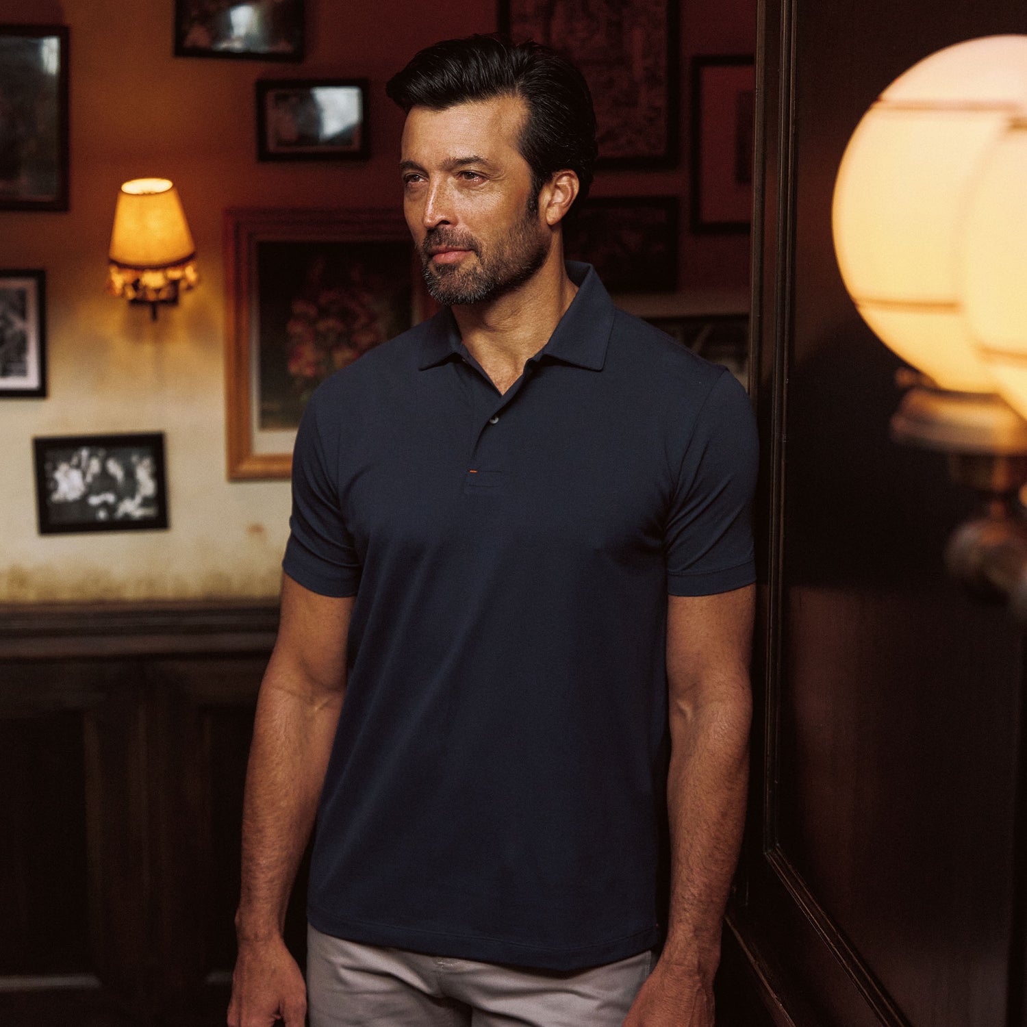 Man wears Tom Beckbe Hutton Performance Polo indoors near a wall with framed photos and warm lighting.
