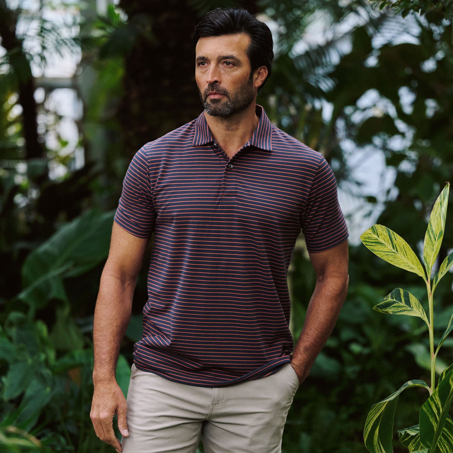 A man in the Tom Beckbe Hutton Performance Polo and beige shorts stands outdoors among green plants with a neutral expression.