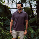 A man wears the Tom Beckbe Hutton Performance Polo and khaki shorts, standing among tropical plants.
