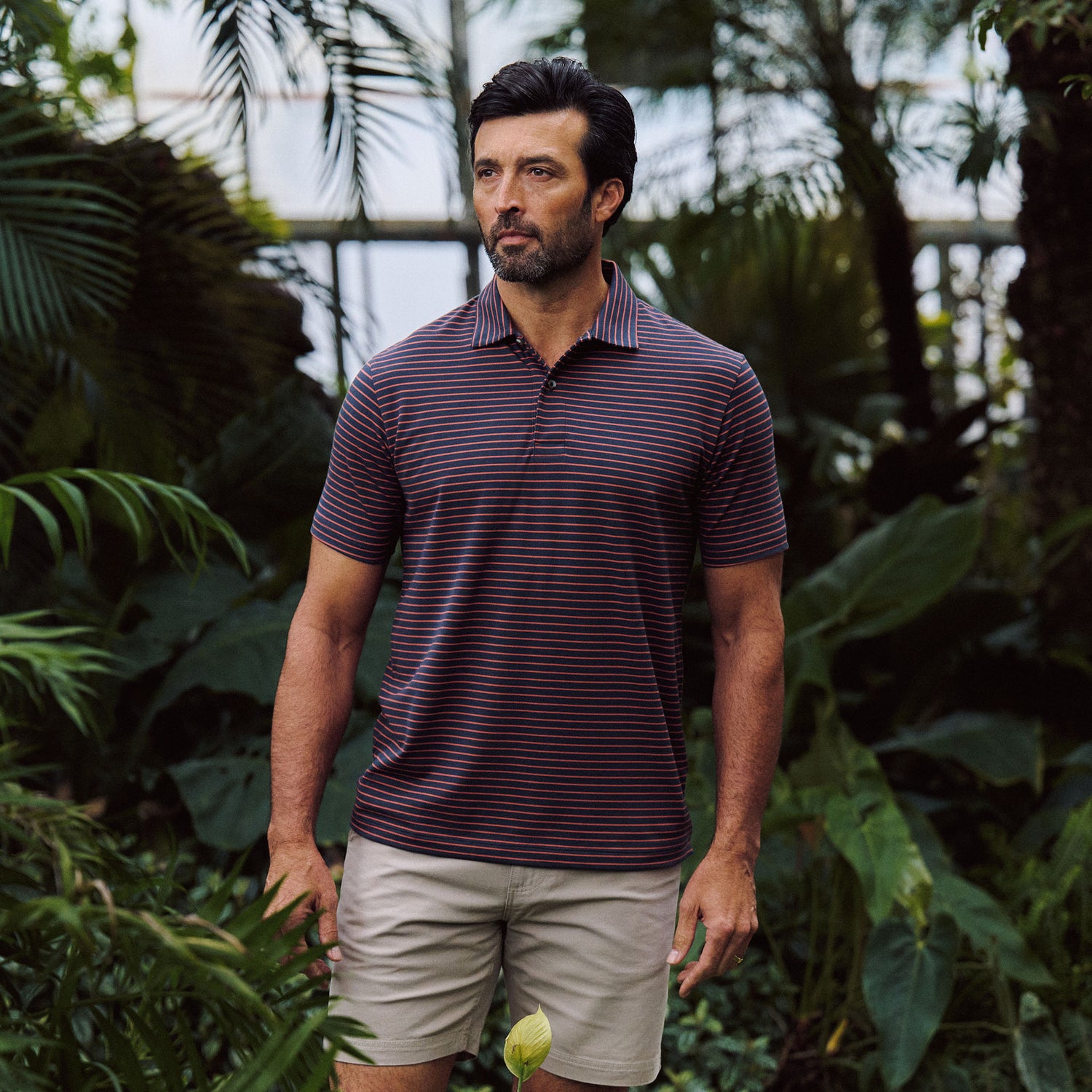 A man wears the Tom Beckbe Hutton Performance Polo and khaki shorts, standing among tropical plants.