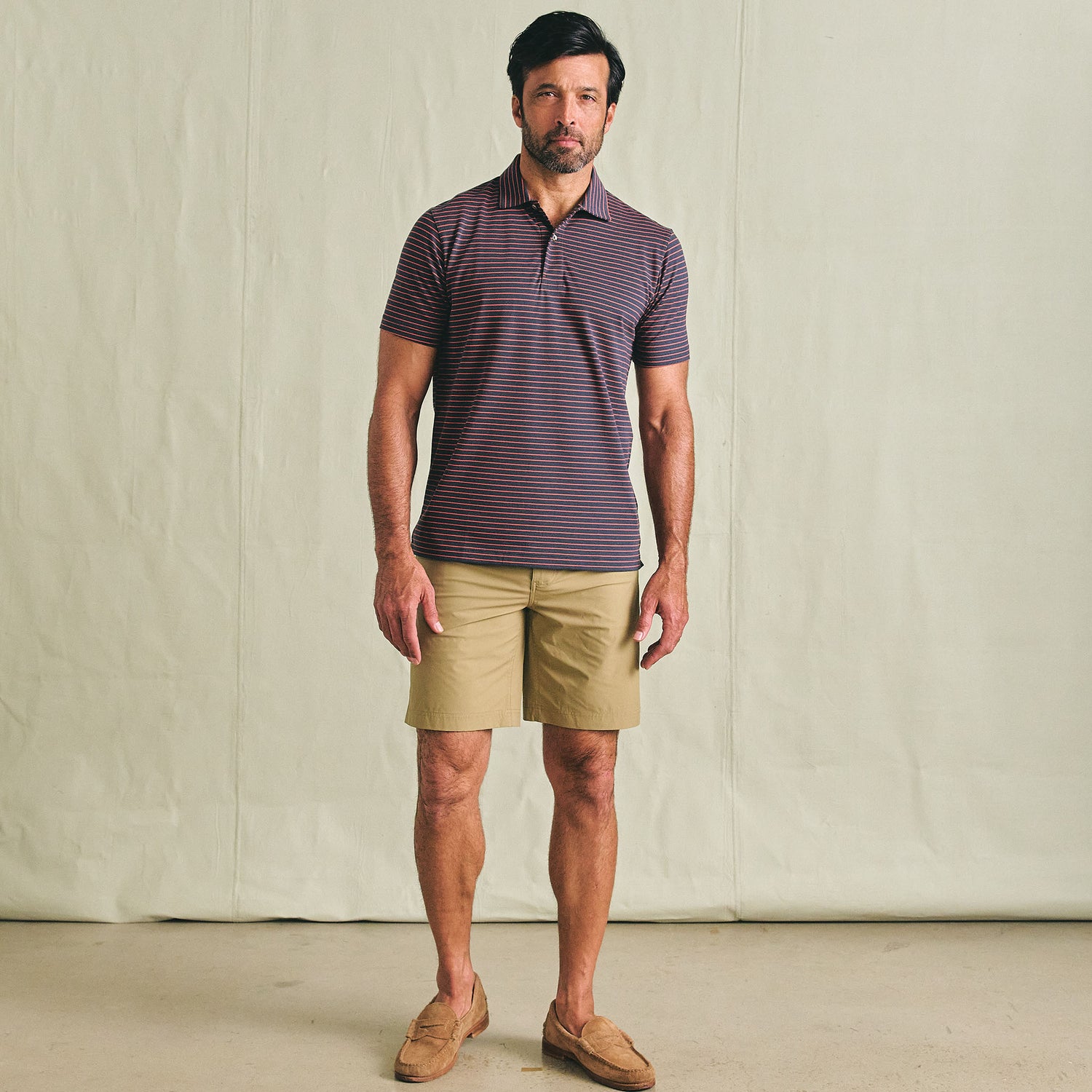 A man wears the Tom Beckbe Hutton Performance Polo, tan shorts, and brown loafers against a plain backdrop.