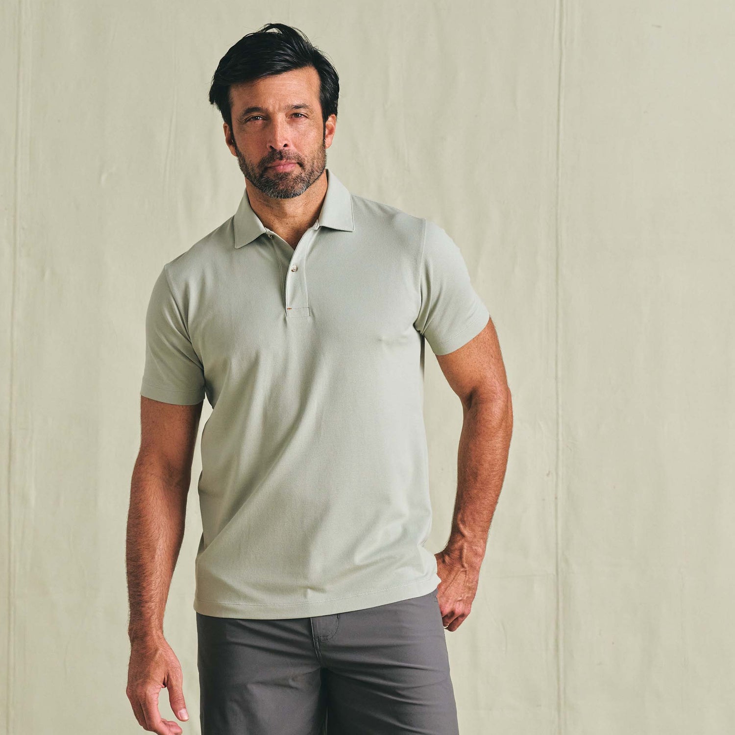 Man wearing a Tom Beckbe Hutton Performance Polo in light gray and dark gray pants against a plain light wall.