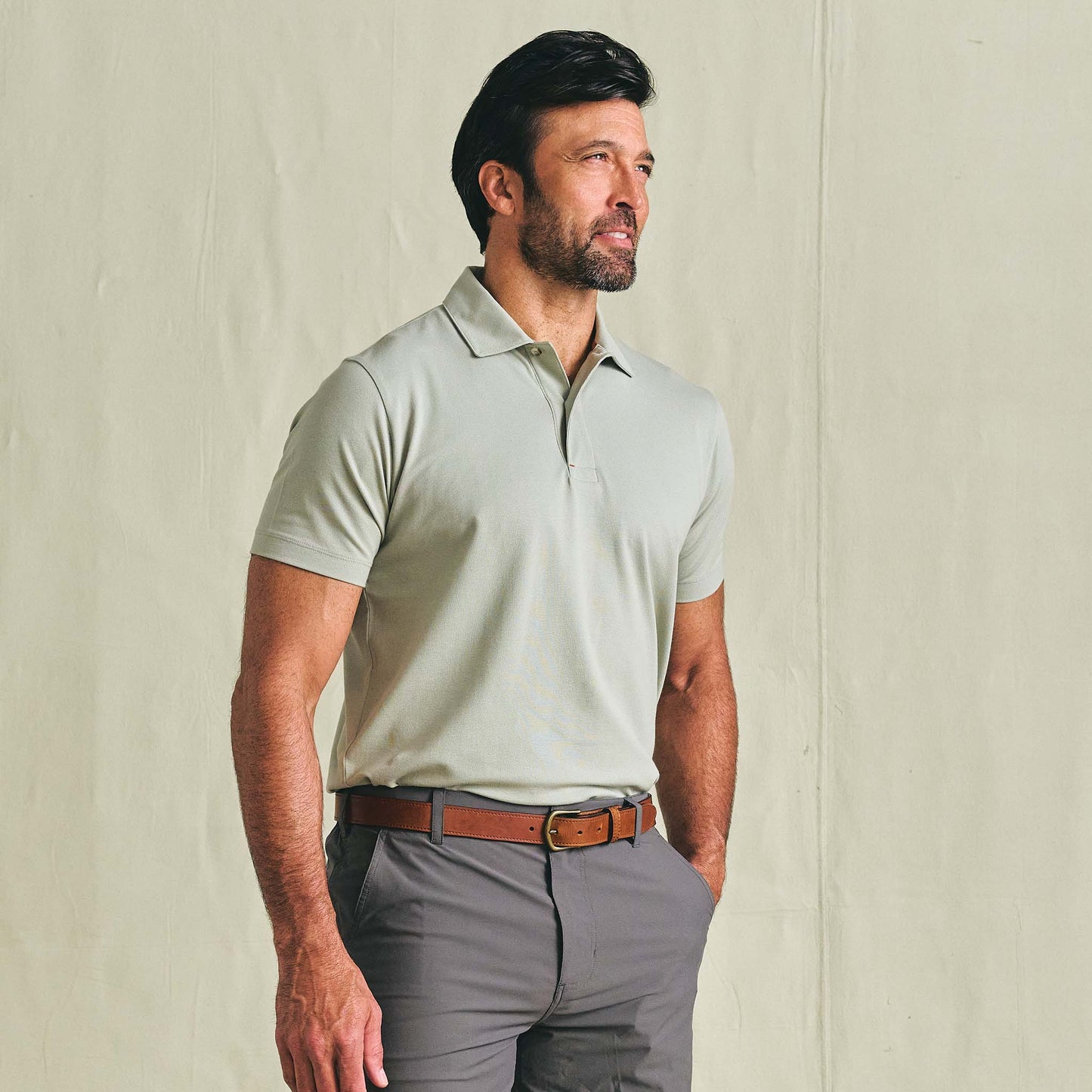 Man wearing the Tom Beckbe Hutton Performance Polo and grey pants with a brown belt against a light background.