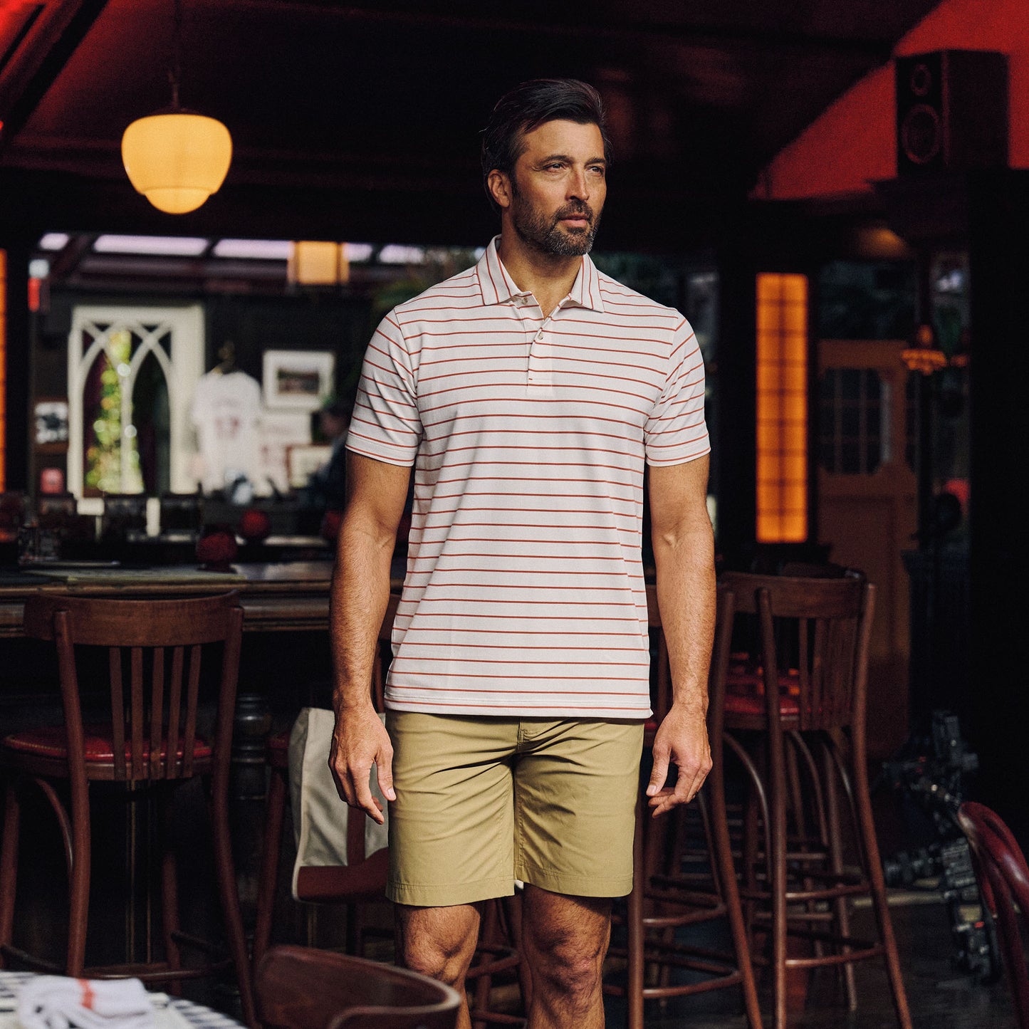 A man in a Tom Beckbe Hutton Performance Polo and khaki shorts stands in a dimly lit bar or restaurant.