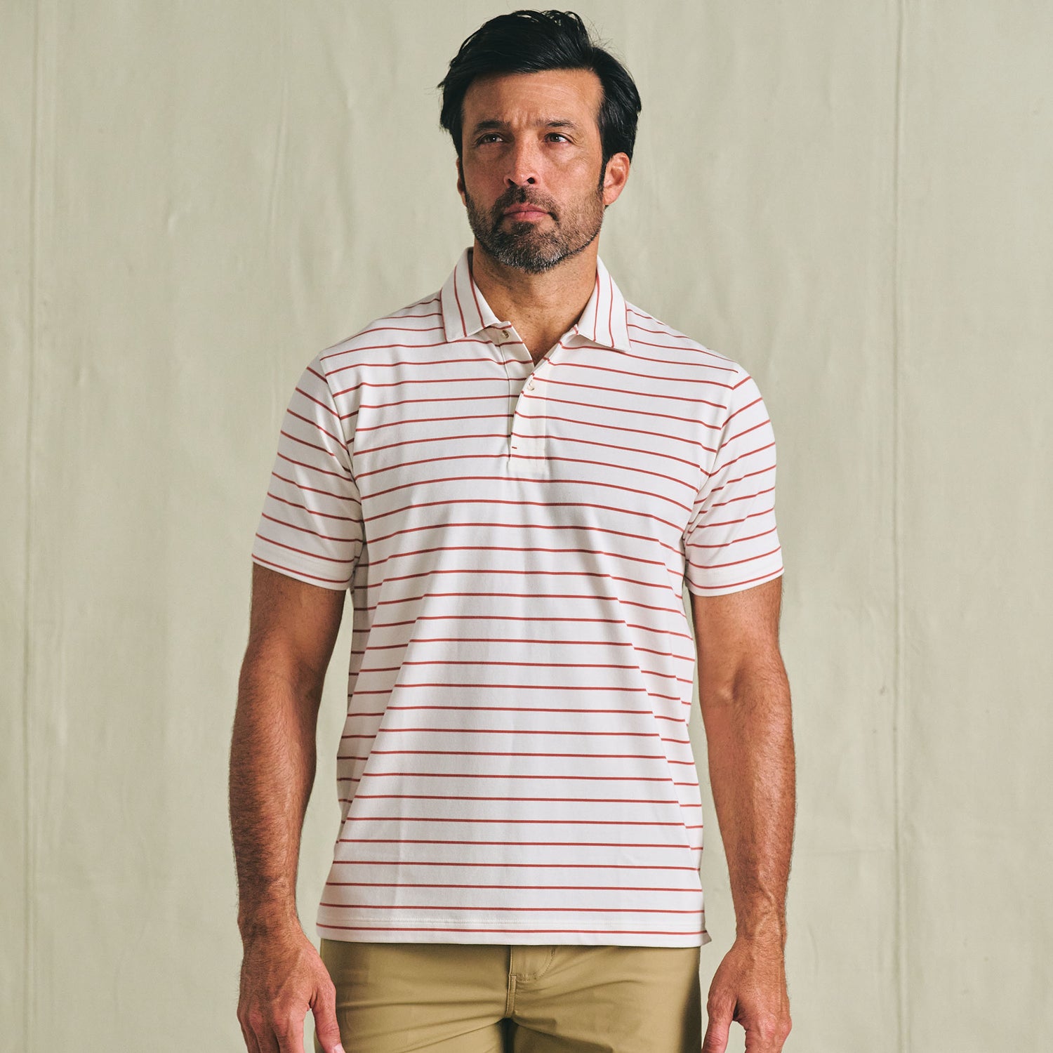 Man wearing a Tom Beckbe Hutton Performance Polo with red stripes and tan pants against a plain background.