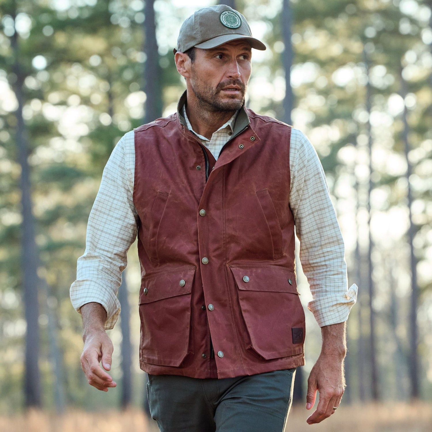 A man in a Fox Red Kinsman Vest by Tom Beckbe walks through a forest, looking to the side.