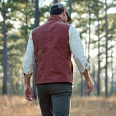 Man walks through forest in Tom Beckbes Fox Red Kinsman Vest and cap, with green pants and a plaid shirt.