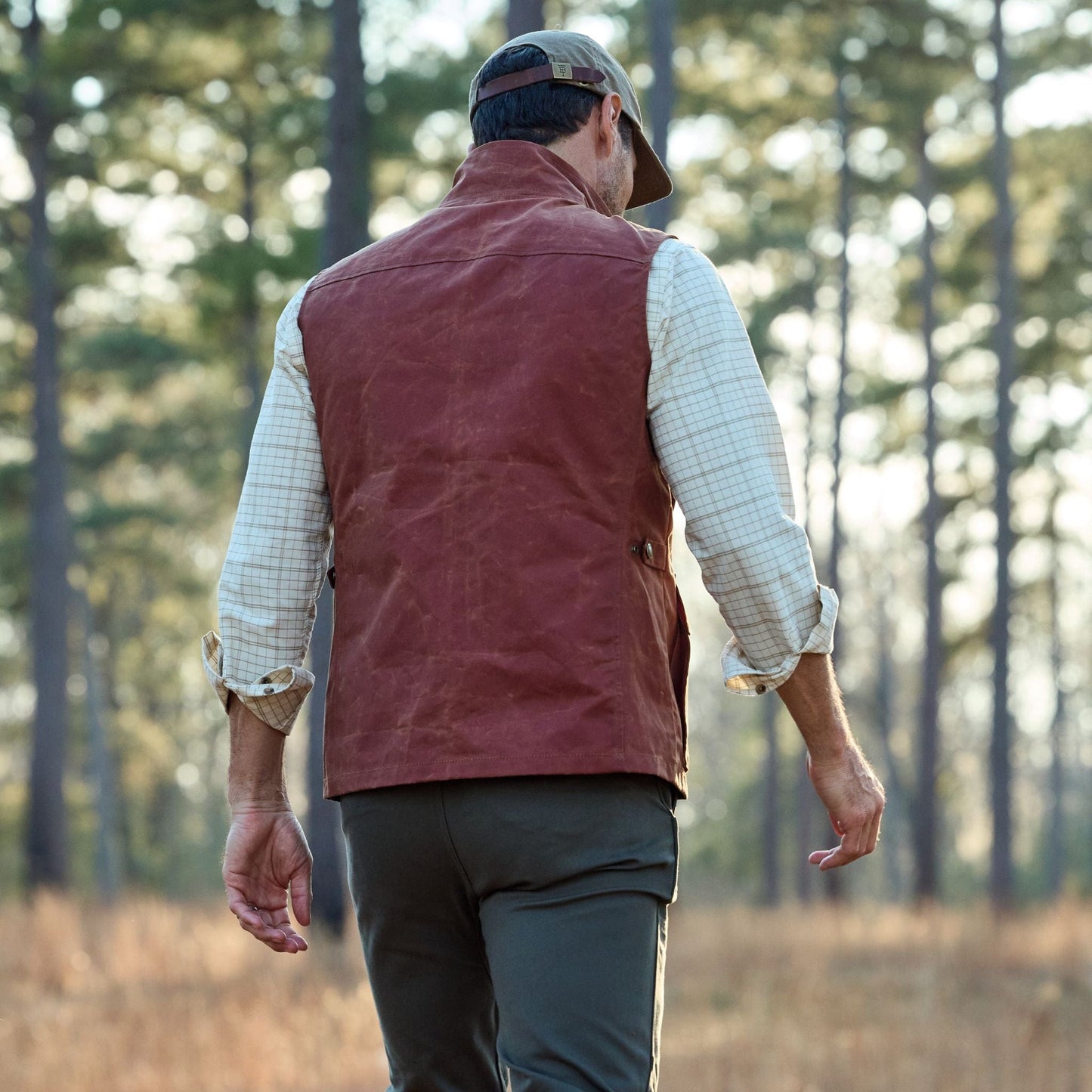 Man walks through forest in Tom Beckbes Fox Red Kinsman Vest and cap, with green pants and a plaid shirt.