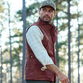 A man in a Tom Beckbe Fox Red Kinsman Vest and cap stands facing trees in a forest.