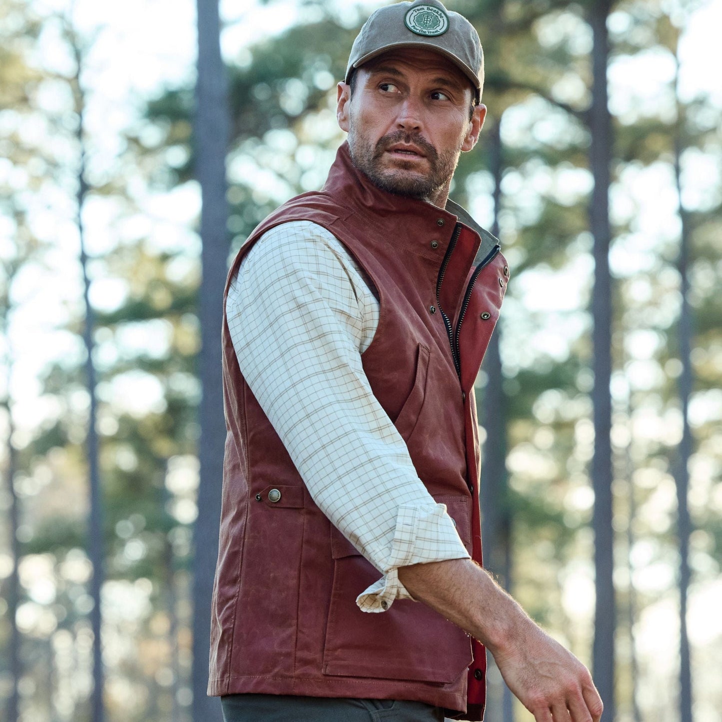 A man in a Tom Beckbe Fox Red Kinsman Vest and cap stands facing trees in a forest.