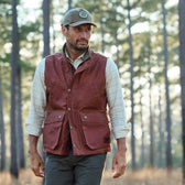 A man in a Tom Beckbe Fox Red Kinsman Vest and a green cap strolls through the forest with sunlight filtering past branches.