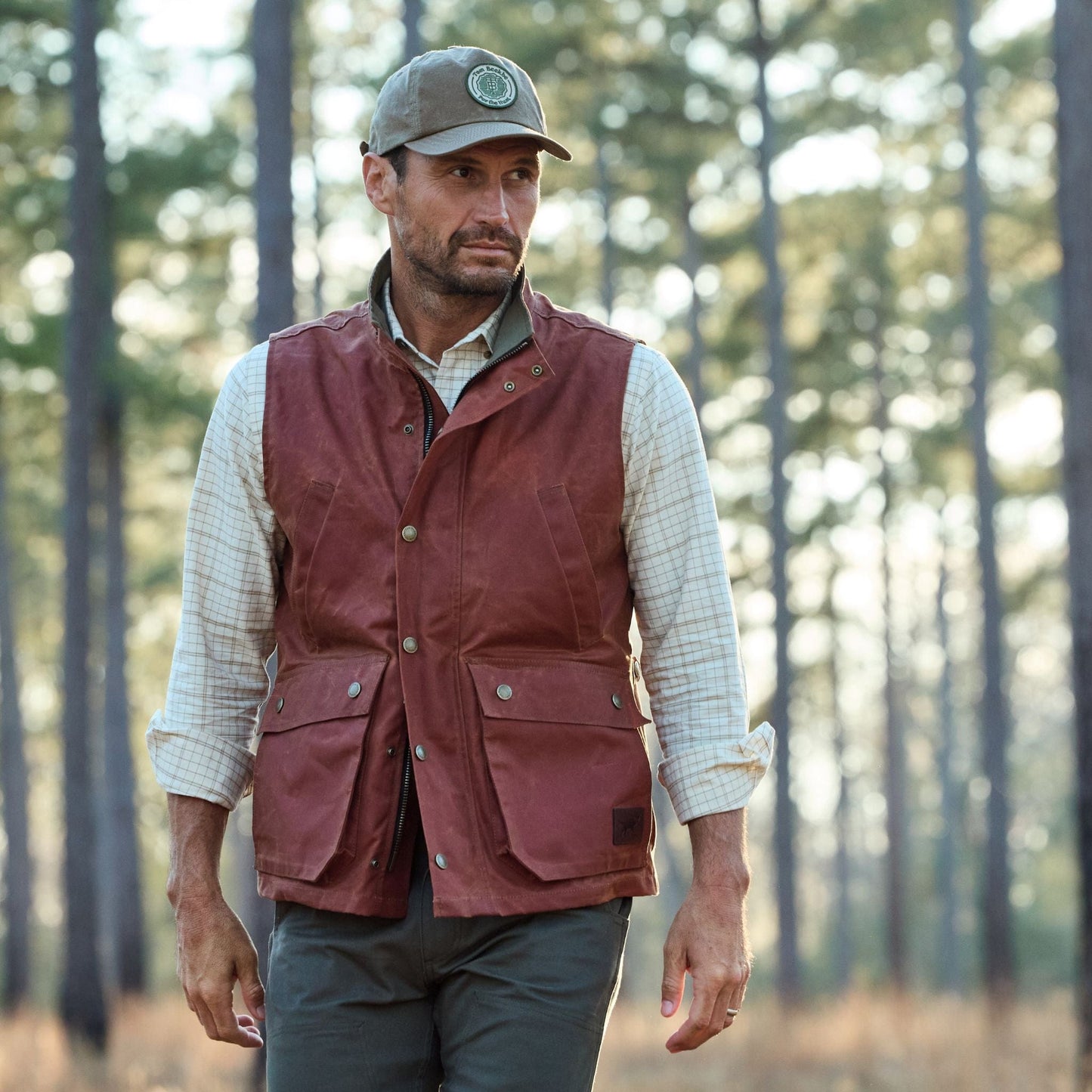 A man in a Tom Beckbe Fox Red Kinsman Vest and a green cap strolls through the forest with sunlight filtering past branches.
