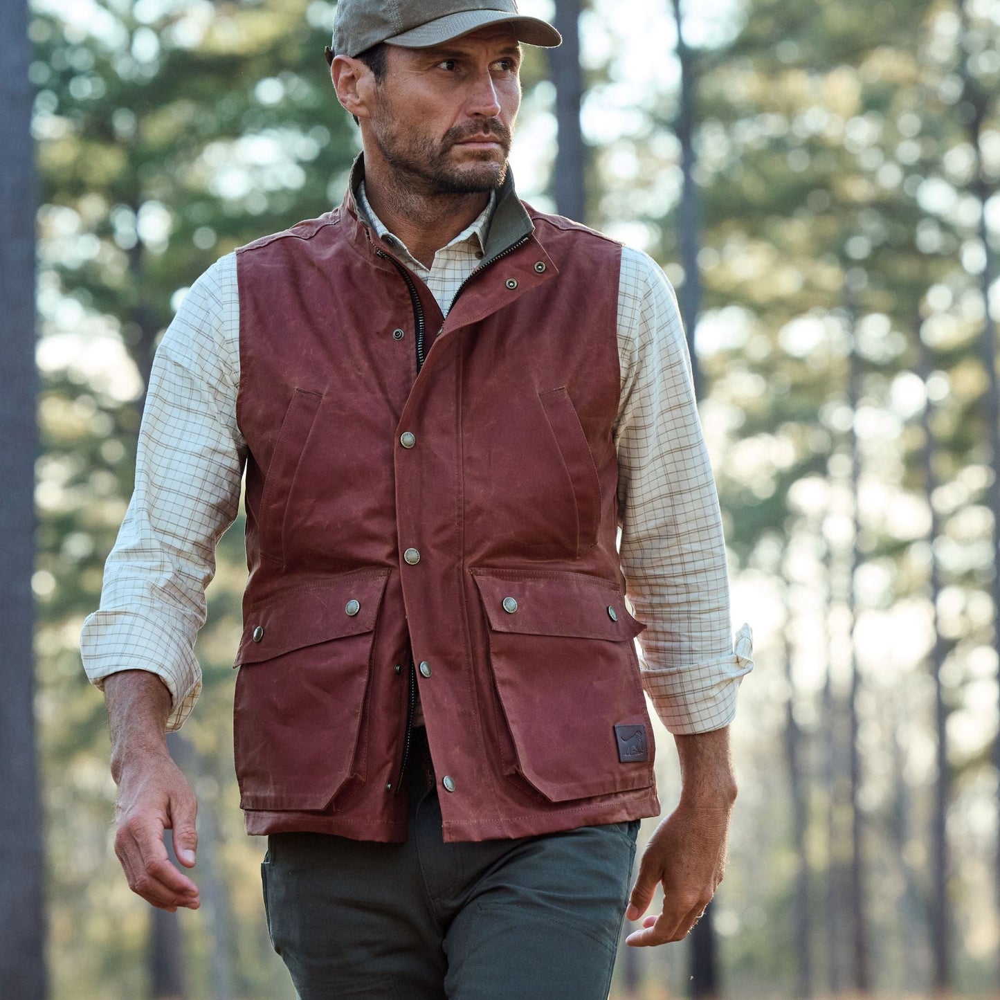 A man in a Tom Beckbe Fox Red Kinsman Vest, plaid shirt, and cap walks through a forest with trees behind him.