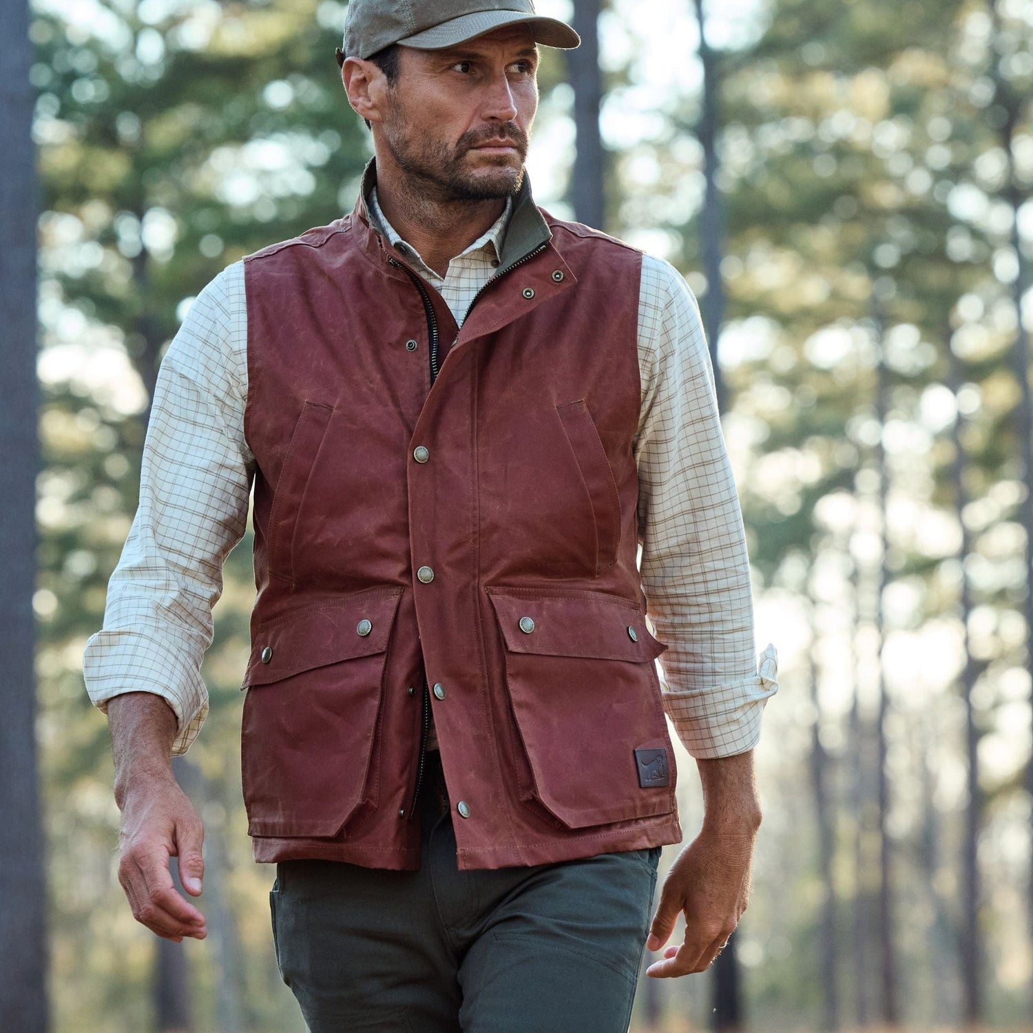 A man in a Tom Beckbe Fox Red Kinsman Vest, plaid shirt, and cap walks through a forest with trees behind him.