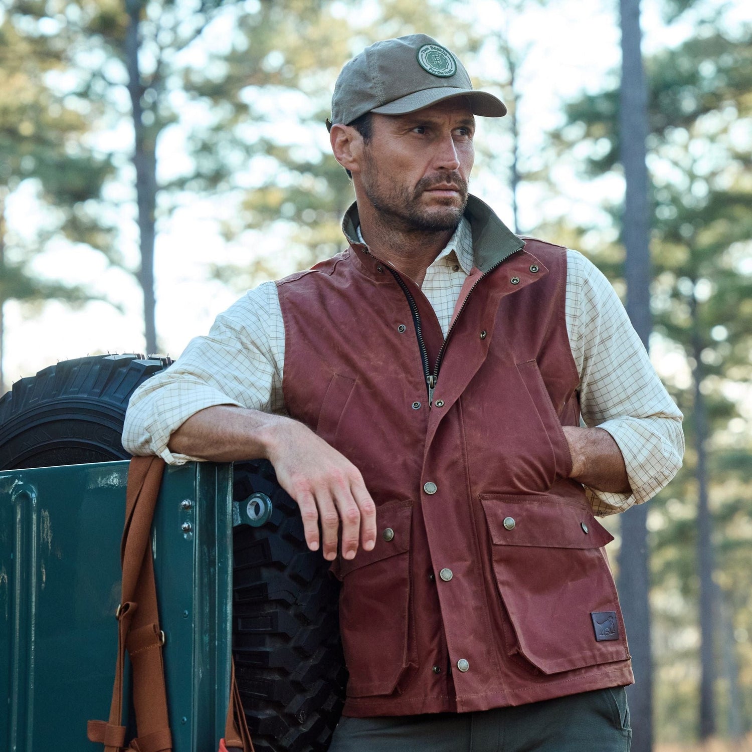 A man in a Fox Red Kinsman Vest by Tom Beckbe and cap leans on a vehicle with spare tire in a forest setting.