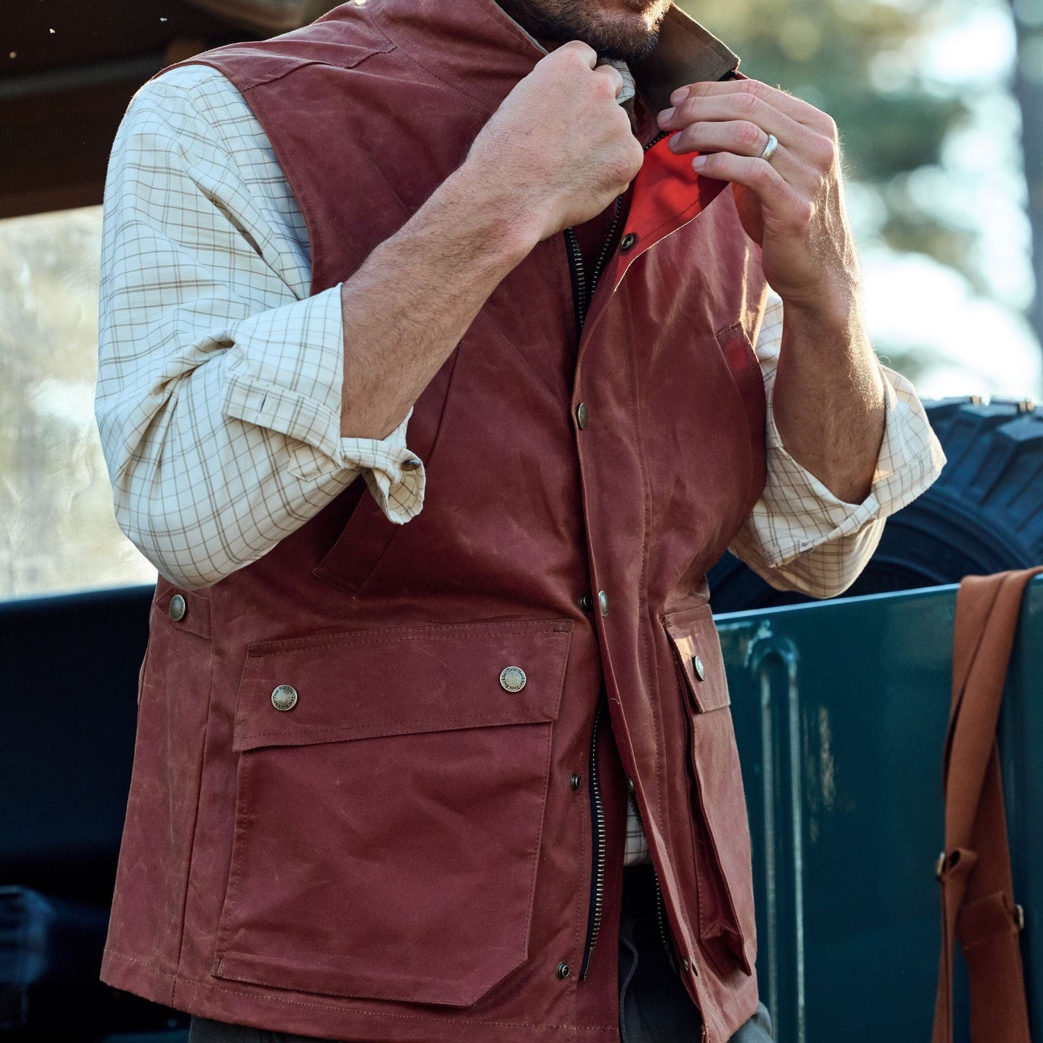 A person in a limited edition Tom Beckbe Fox Red Kinsman Vest over a plaid shirt adjusts the collar near a vehicle.