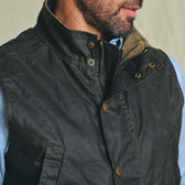 Man wears Tom Beckbe Kinsman Vest—dark waxed canvas, collar up—over a light blue shirt, partially unzipped.
