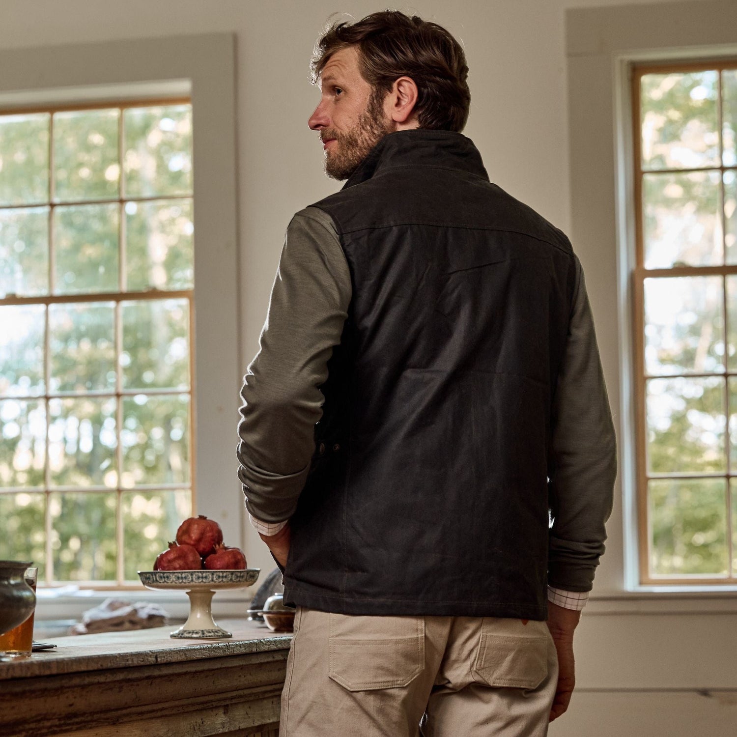 Wearing a Kinsman Vest by Tom Beckbe, a man stands by large windows with a bowl of apples on the counter beside him.
