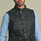 A man wears the Tom Beckbe Kinsman Vest, a black weatherproof shell, over a light blue collared shirt.