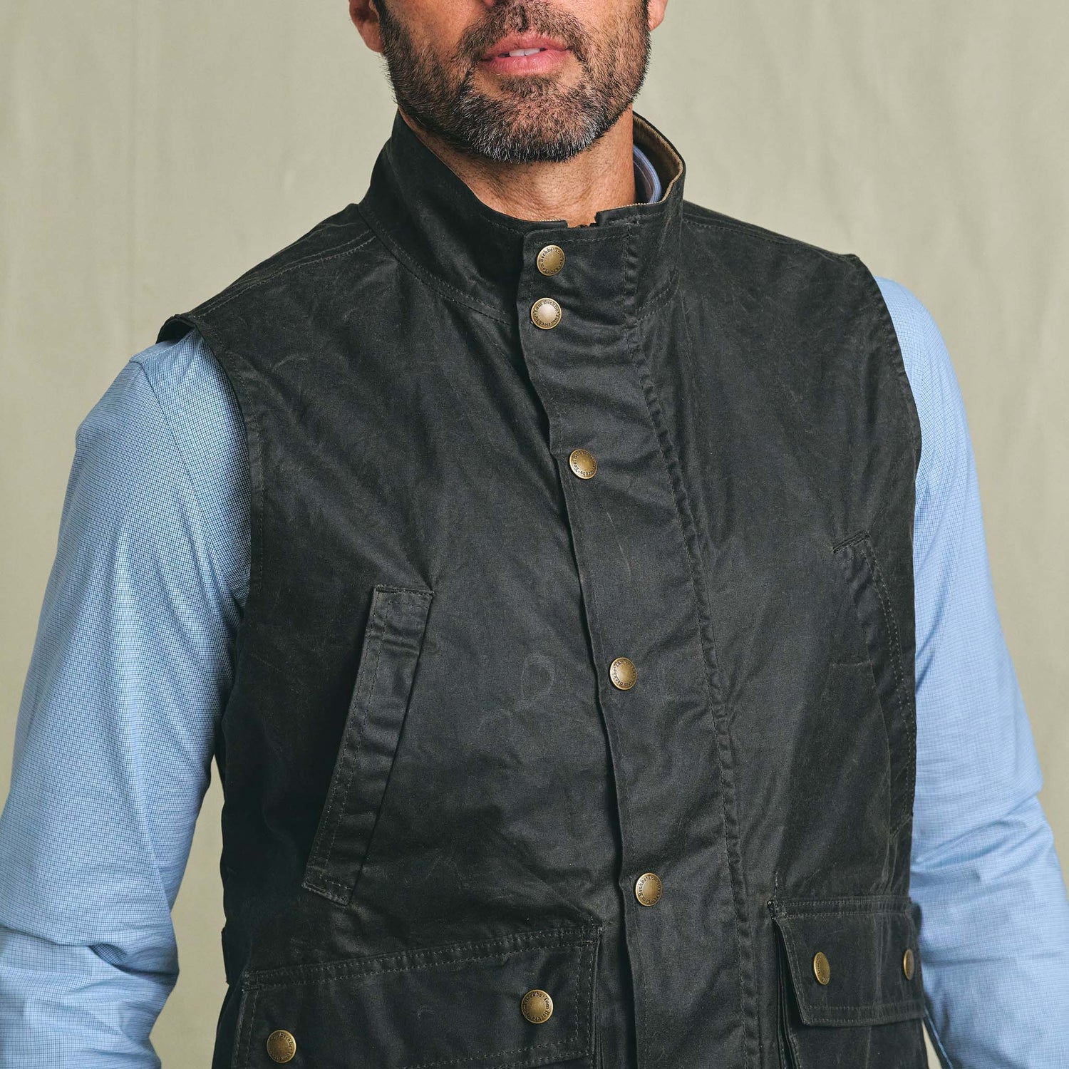 A man wears the Tom Beckbe Kinsman Vest, a black weatherproof shell, over a light blue collared shirt.