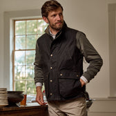 Man wearing a Tom Beckbe Kinsman Vest and khaki pants stands near a window, dishes on wooden table.