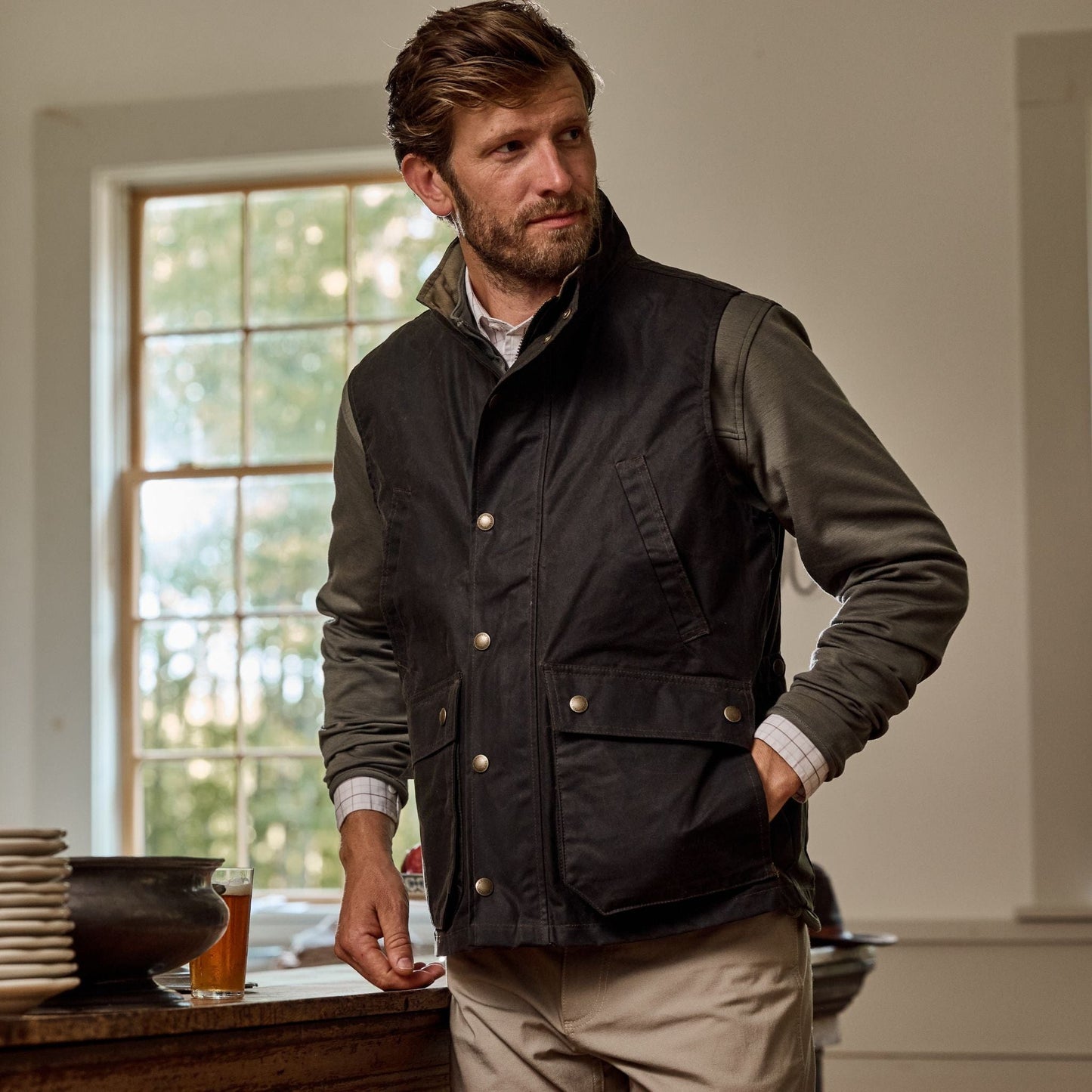 Man wearing a Tom Beckbe Kinsman Vest and khaki pants stands near a window, dishes on wooden table.
