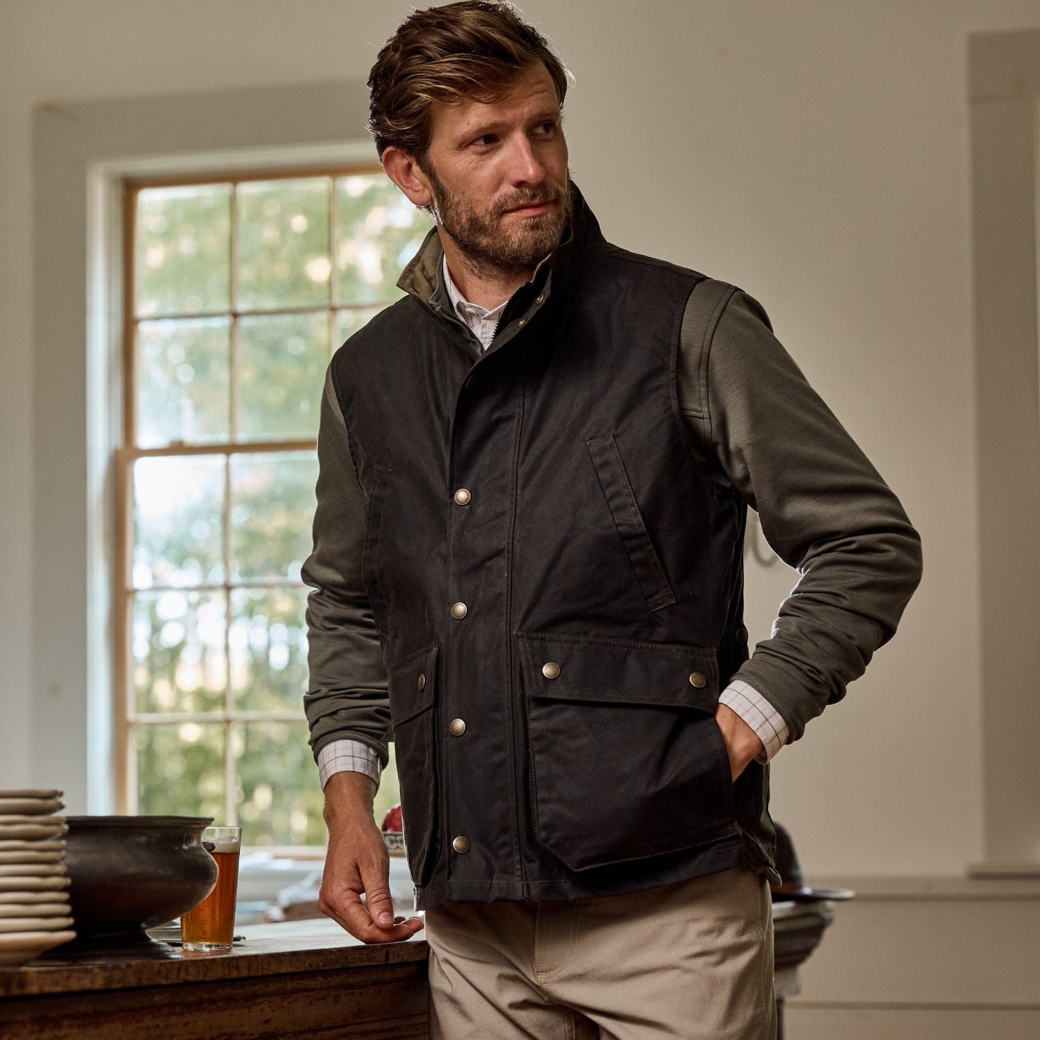 Man wearing a Tom Beckbe Kinsman Vest and khaki pants stands near a window, dishes on wooden table.