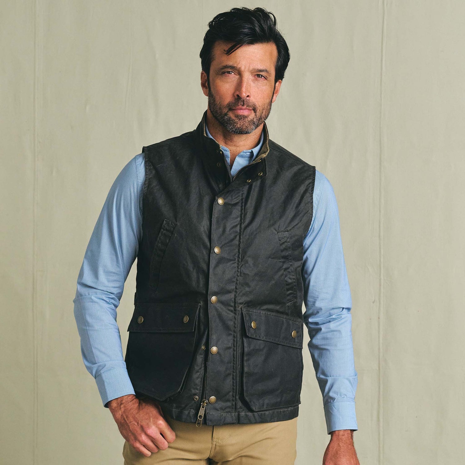 Man wears the Tom Beckbe Kinsman Vest over a blue shirt and khakis, standing before a plain beige background.