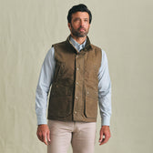 A man wears the Tom Beckbe Kinsman Vest in brown over a light blue shirt and beige pants against a beige background.