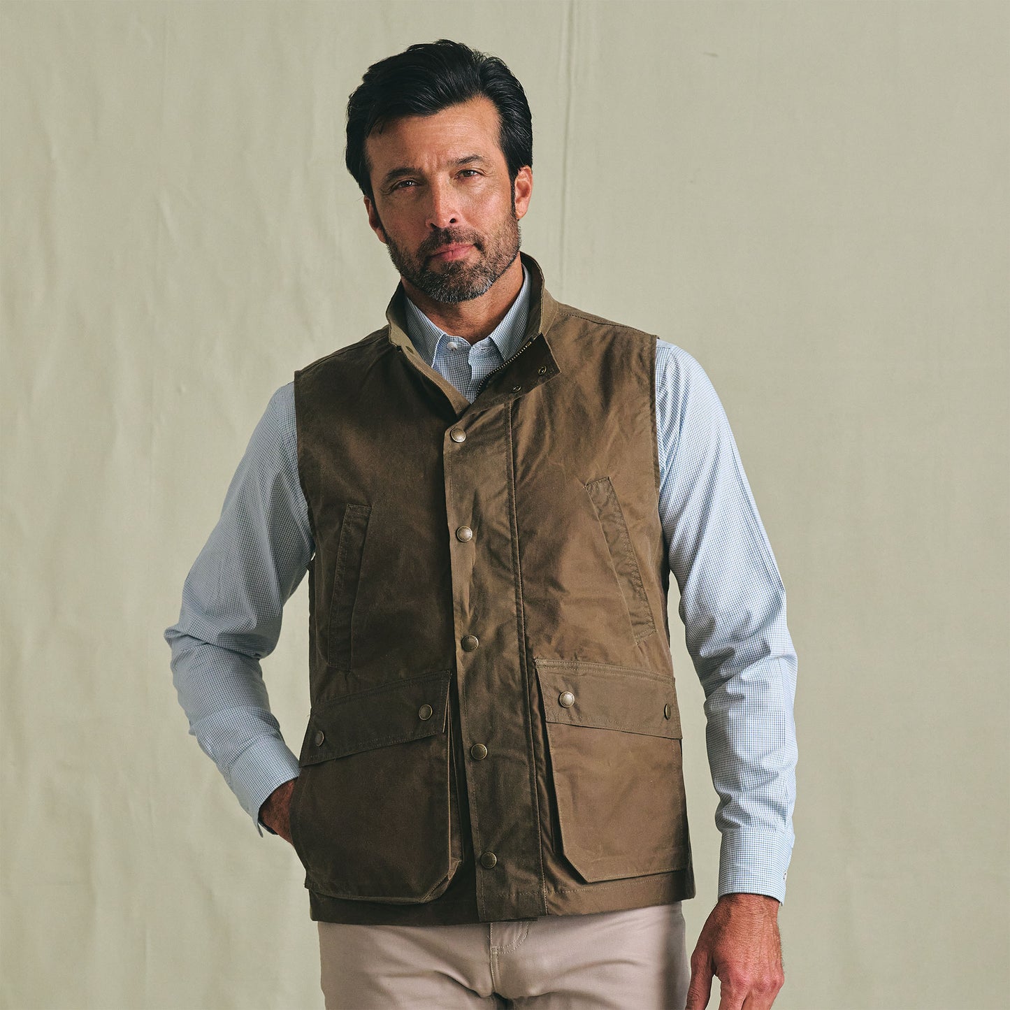 A man wears the Tom Beckbe Kinsman Vest over a light blue shirt, standing against a plain beige background.