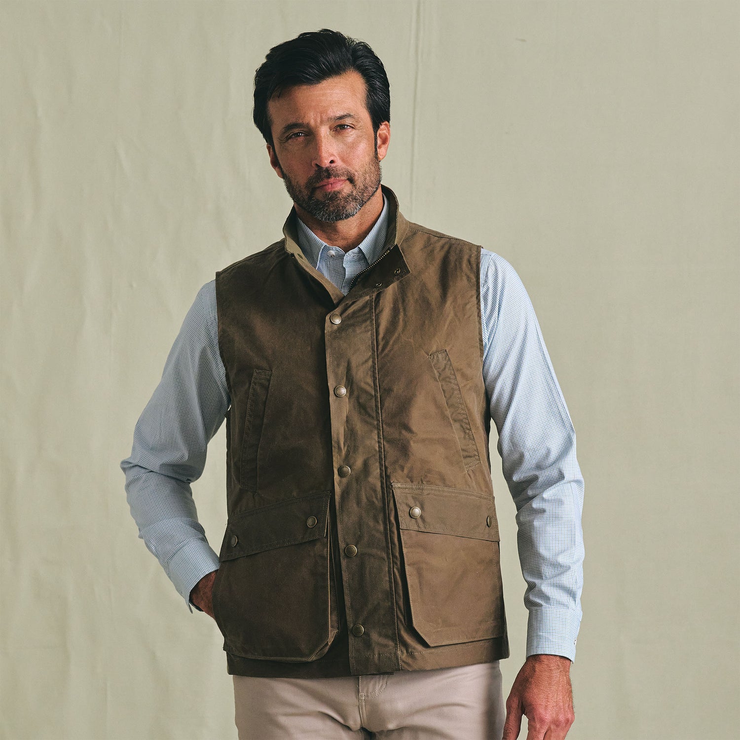 A man wears the Tom Beckbe Kinsman Vest over a light blue shirt, standing against a plain beige background.