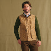 Man wears Tom Beckbe Kinsman Vest over green sweater, brown pants; stands before beige fabric backdrop.
