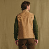 Man faces away, wearing the Tom Beckbe Kinsman Vest over a green sweater and brown pants against a beige fabric backdrop.