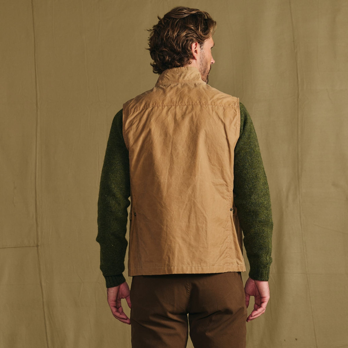 Man faces away, wearing the Tom Beckbe Kinsman Vest over a green sweater and brown pants against a beige fabric backdrop.