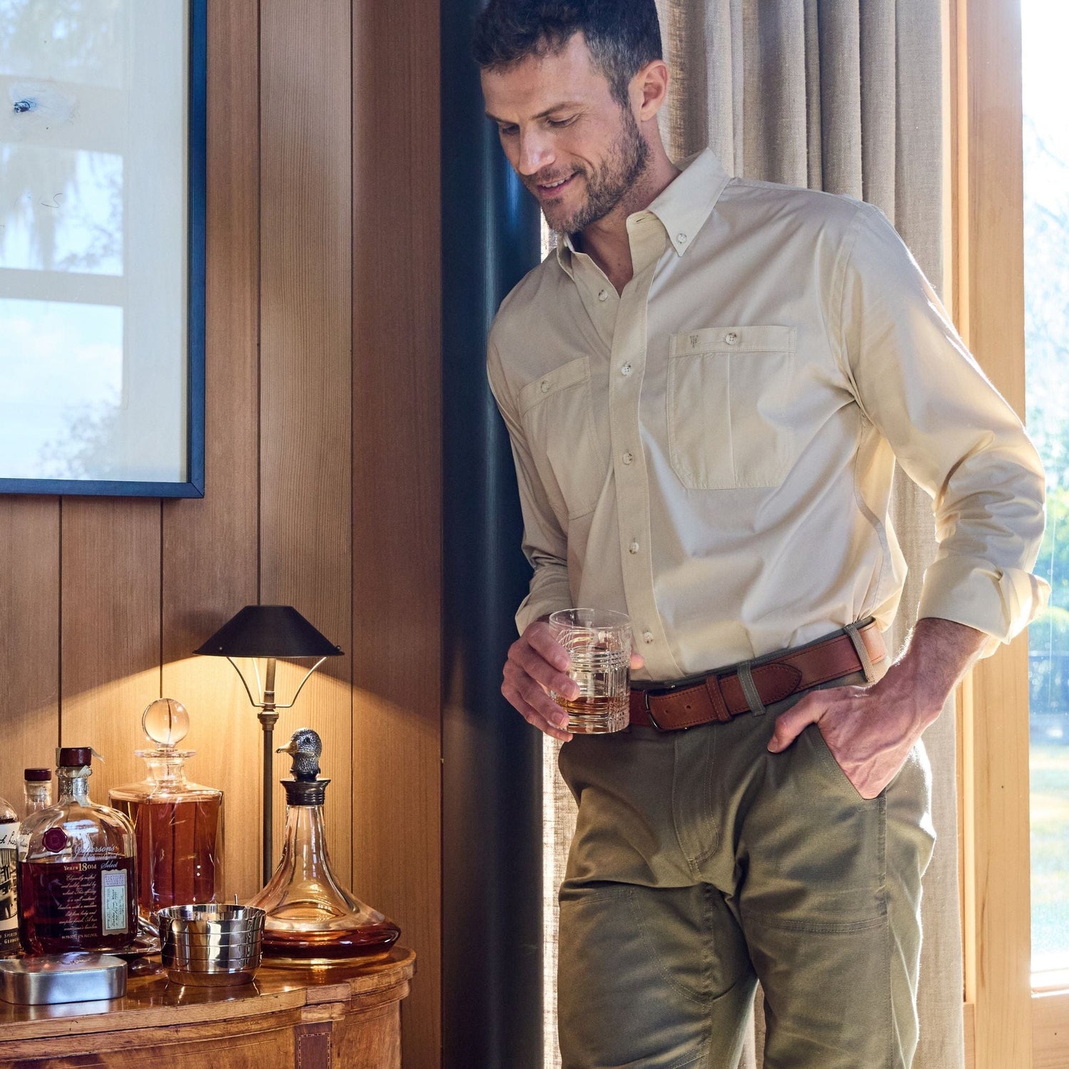 A man in a beige shirt and a Tom Beckbe leather belt holds a drink by a wooden cabinet with liquor bottles and a lamp.