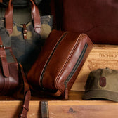 Tom Beckbe Leather Dopp Kit, green camo backpack, and baseball cap rest on a wooden surface.